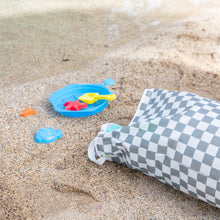 Load image into Gallery viewer, A beach scene with a Bumkins Extra Large Wet Bag: Charcoal Check, a blue bucket, yellow shovel, and colorful toys on sand by the water's edge.