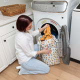 A person kneels by a front-load washer, placing colorful clothes into a Bumkins Extra Large Wet Bag: Winnie + Friends.