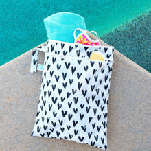 Load image into Gallery viewer, Bumkins Wet/Dry Bag: Hearts sits near a pool, filled with a towel, swimsuit, and sunscreen.