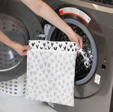 Load image into Gallery viewer, A person holds Bumkins Wet/Dry Bag: Hearts in front of an open front-loading washing machine.