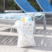 Load image into Gallery viewer, Bumkins Wet/Dry Bag: Palm Daze sits in front of empty lounge chairs, with a water bottle, towel, and swim gear inside.