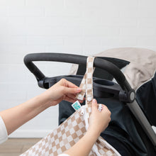 Load image into Gallery viewer, Hands attach a Bumkins Wet/Dry Bag: Sand Check, made of waterproof fabric, to a stroller using snap buttons on the strap.