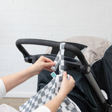 Hands attach the Bumkins Wet/Dry Bag: Charcoal Check to a stroller handle against a white brick wall.