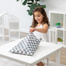 Load image into Gallery viewer, A young girl in a white dress sits at a white table, happily opening her Bumkins Wet/Dry Bag: Charcoal Check in a bright room.