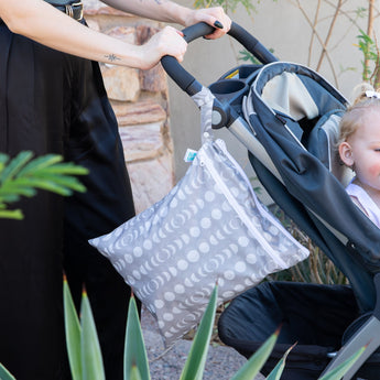 A person pushes a stroller with a child. A Bumkins Wet/Dry Bag: Lunar Phase hangs from the handle, ideal for travel.