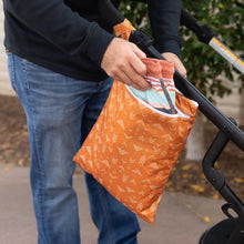 Load image into Gallery viewer, A person places clothes into a Bumkins Wet/Dry Bag: Grounded, attached to a stroller outdoors.