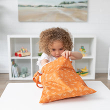 Load image into Gallery viewer, A curly-haired child peers into a Bumkins Wet/Dry Bag: Grounded at a white table, surrounded by toy-filled shelves.