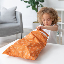 Load image into Gallery viewer, A curly-haired child opens a Bumkins Wet/Dry Bag: Grounded at a white table. A sofa and plant add charm to the background.