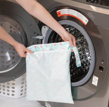 Load image into Gallery viewer, A person holds Bumkins Wet/Dry Bag: Rainbows in front of an open front-loading washing machine.