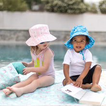 Load image into Gallery viewer, Two kids in sun hats sit by a pool on a colorful mat. One holds a book, while the other grabs a towel from a Bumkins Wet/Dry Bag: Rainbows.