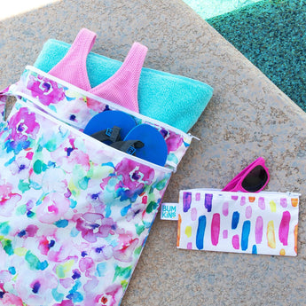 Bumkins Wet/Dry Bag: Watercolor lays next to a pool, holding flip flops, a swimsuit, and a towel; a small bag with sunglasses sits nearby.