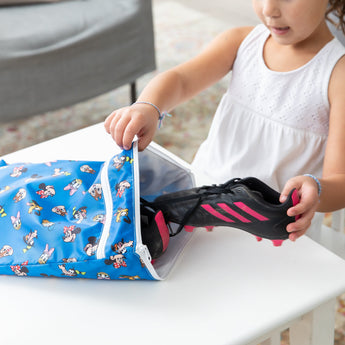 A child places a soccer cleat into a blue Bumkins Wet/Dry Bag: Mickey & Friends Forever on a white table.