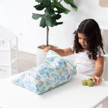 Load image into Gallery viewer, A young girl opens a Bumkins Wet/Dry Bag: Aloha Stitch while holding a green toy at a white table, with a potted plant in the background.