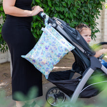 Load image into Gallery viewer, A person pushes a stroller with a Bumkins Wet/Dry Bag: Aloha Stitch hanging from the handle next to green foliage.