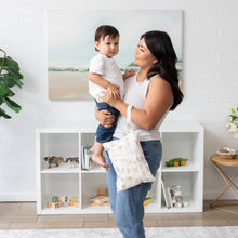 Load image into Gallery viewer, A woman smiles while holding a child and a Bumkins Wet/Dry Clutch: Palm Daze in a bright living room in front of shelves filled with toys.