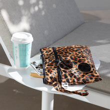 Load image into Gallery viewer, A Roamio by Bumkins Wet/Dry Clutch: Leopard with sunglasses & a camera sits on a white table with a coffee cup by a gray chair outdoors.