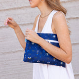 A person in white holds a Bumkins Wet/Dry Clutch: Evil Eye in front of a beige textured wall.