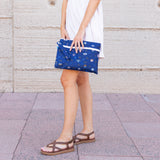 A person in a white dress & sandals holds a Bumkins Wet/Dry Clutch: Evil Eye on tiled pavement against a beige textured wall.
