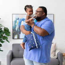 Load image into Gallery viewer, A smiling man in a blue shirt holds a young child and a Bumkins Wet/Dry Clutch: Evil Eye in a modern living room with artwork.