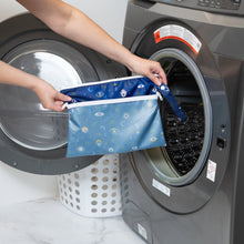 Load image into Gallery viewer, A person holds Bumkins Wet/Dry Clutch: Evil Eye in front of a front-loading washing machine.