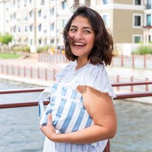 Load image into Gallery viewer, A smiling woman with short brown hair holds a Roamio by Bumkins Wet/Dry Clutch: Nautical Stripe with beige buildings in the background.