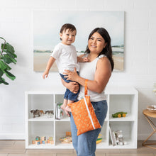Load image into Gallery viewer, A woman holds a toddler and smiles beside a white brick wall, bookshelf, and plant, holding a Bumkins Wet/Dry Clutch: Grounded on her arm.