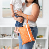 A person in white tank top holds a child and a Bumkins Wet/Dry Clutch: Grounded in a bright room with a bookshelf filled with toys behind.