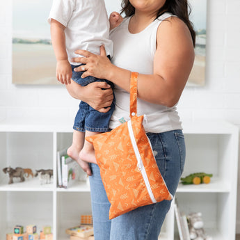 A person in white tank top holds a child and a Bumkins Wet/Dry Clutch: Grounded in a bright room with a bookshelf filled with toys behind.