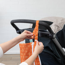 Load image into Gallery viewer, Hands attach the Bumkins Wet/Dry Clutch: Grounded, an orange patterned waterproof bag, to a stroller against a white brick wall.