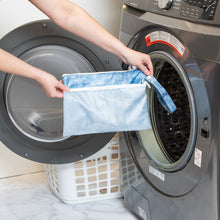Load image into Gallery viewer, A person holds Roamio by Bumkins Wet/Dry Clutch: Crushed Denim in front of an open front-loading washing machine.