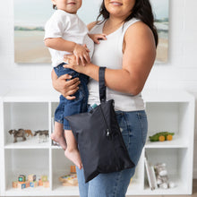 Load image into Gallery viewer, A woman in a toy-filled living room holds a toddler and carries a Bumkins Wet/Dry Clutch: Black, featuring waterproof fabric.