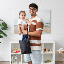 Load image into Gallery viewer, A man with a toddler smiles at the camera in a toy-filled room, holding Bumkins Wet/Dry Clutch: Black on his arm.