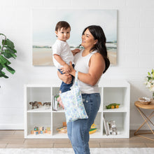Load image into Gallery viewer, A woman cradles her baby near a toy-filled bookshelf; a Bumkins Wet/Dry Clutch: Aloha Stitch hangs from her arm.