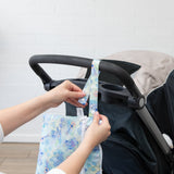A person attaches a Bumkins Wet/Dry Clutch: Aloha Stitch to a stroller handle against a white brick wall.