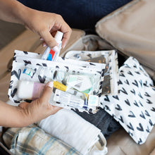 Load image into Gallery viewer, Hands fill a Bumkins Clear Travel Bag 3-Pack: Hearts with toiletries above a packed suitcase, with the other bags on top.