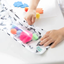 Load image into Gallery viewer, Close-up of a child's hand zipping a Bumkins Clear Travel Bag 3-Pack: Hearts filled with colorful pom-poms on a white table.
