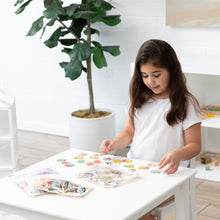 Load image into Gallery viewer, A girl in a white shirt assembles a colorful puzzle at a table, surrounded by Bumkins Clear Travel Bag 3 Pack: Palm Daze and a potted plant.