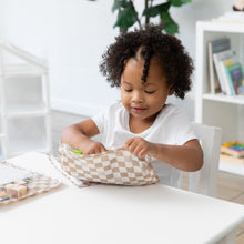 Load image into Gallery viewer, A curly-haired child eagerly unzips a Roamio by Bumkins Clear Travel Bag 3 Pack: Sand Check at a white table, surrounded by books & a plant.