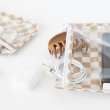 Load image into Gallery viewer, Close-up of a Roamio by Bumkins Clear Travel Bag 3 Pack: Sand Check with a hair clip & coiled cord on a white background near an earbud case.