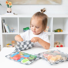 Load image into Gallery viewer, A toddler with a ponytail reaches into a Bumkins Clear Travel Bag 3 Pack: Charcoal Check at a table with colorful toys.