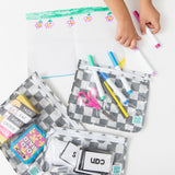 A child draws with markers as Bumkins Clear Travel Bag 3 Pack: Charcoal Check holds scissors, cards, and supplies on a white table.
