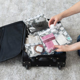 Hands placing Bumkins Clear Travel Bag 3 Pack: Charcoal Check into an open black suitcase on a carpeted floor.