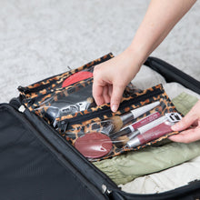 Load image into Gallery viewer, A person packs Roamio by Bumkins Clear Travel Bag 3 Pack: Leopard with makeup into an open suitcase with folded clothes on a gray carpet.