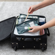 Load image into Gallery viewer, A person places toiletries into a Bumkins Clear Travel Bag 3 Pack: Starry Cactus in an open suitcase on a gray carpet.