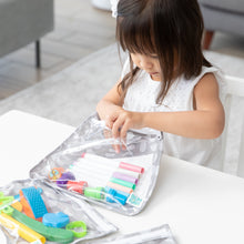 Load image into Gallery viewer, A child in a white dress opens a Bumkins Clear Travel Bag 3 Pack: Lunar Phase filled with colorful markers and scissors at a white table.