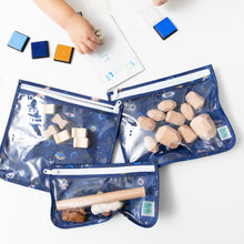 Load image into Gallery viewer, A child's hand plays with stamps beside Bumkins Clear Travel Bag 3 Pack: Evil Eye, filled with wooden toys on a white table.