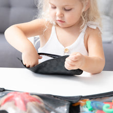 Load image into Gallery viewer, A blonde girl reaches into a Bumkins Clear Travel Bag 3 Pack: Rich Black at a white table.
