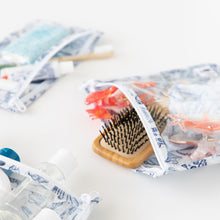 Load image into Gallery viewer, Close-up of Roamio by Bumkins Clear Travel Bag 3 Pack: Shells on a white background with a hairbrush, hair accessories, & toiletries.