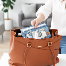 Load image into Gallery viewer, A person places a Roamio by Bumkins Clear Travel Bag 3 Pack: Crushed Denim with a book and cable in a brown purse.