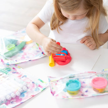A young child sits at a table playing with colorful play dough, surrounded by art supplies in Bumkins Clear Travel Bag 3-Pack: Watercolor.